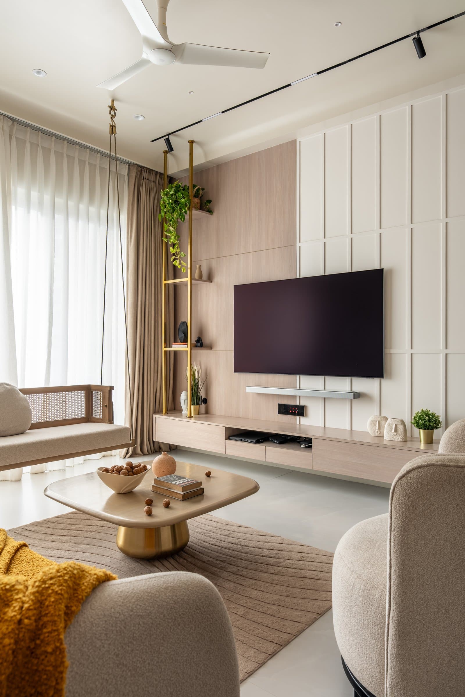 The media wall, where warm wood panelling and cream board-and-batten meet, softened by a brass shelving column threaded with greenery