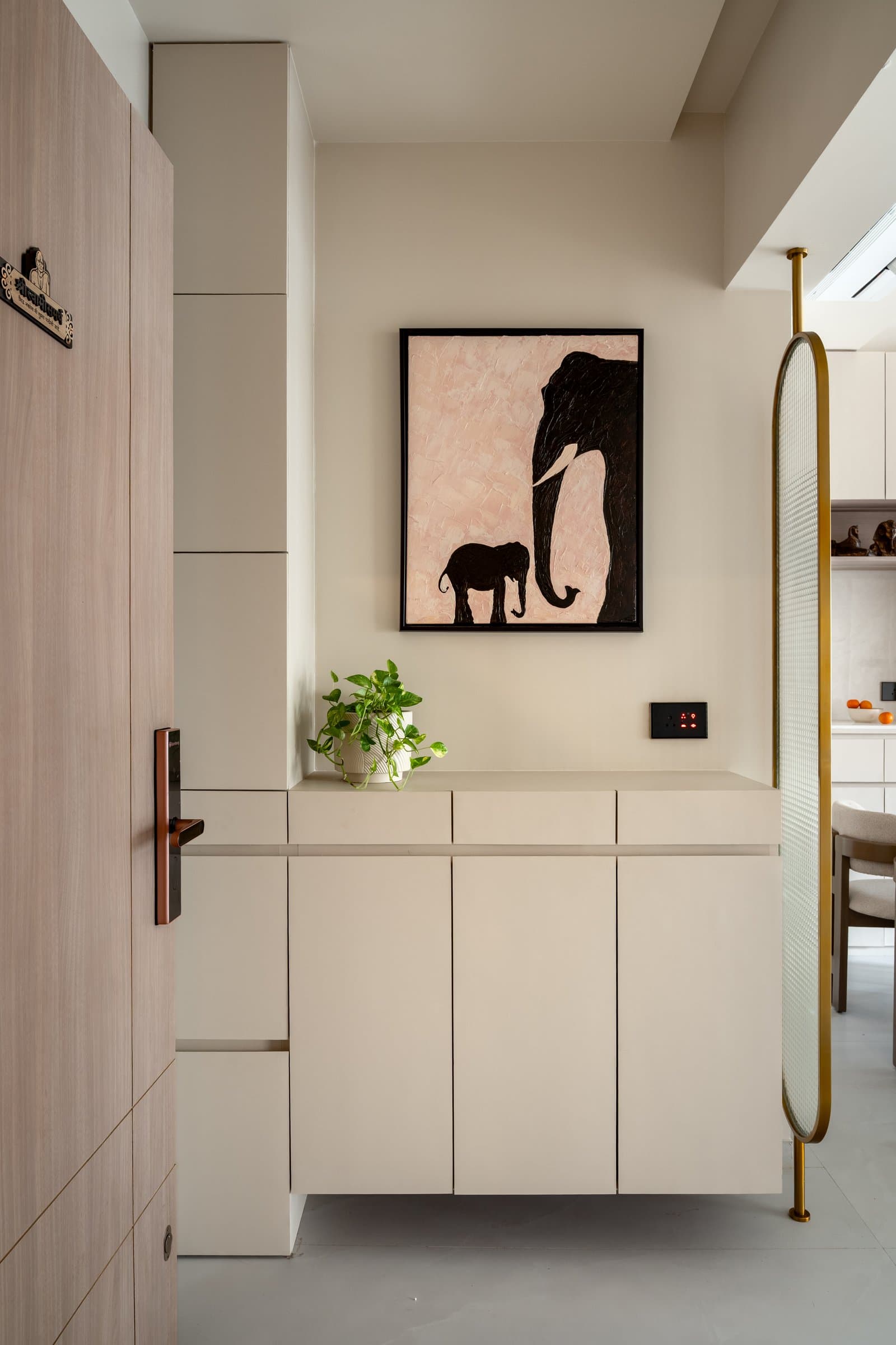 The entry, where a wood-grain door opens onto a concealed storage wall, an elephant artwork, and a brass-framed screen filtering the view to the dining beyond