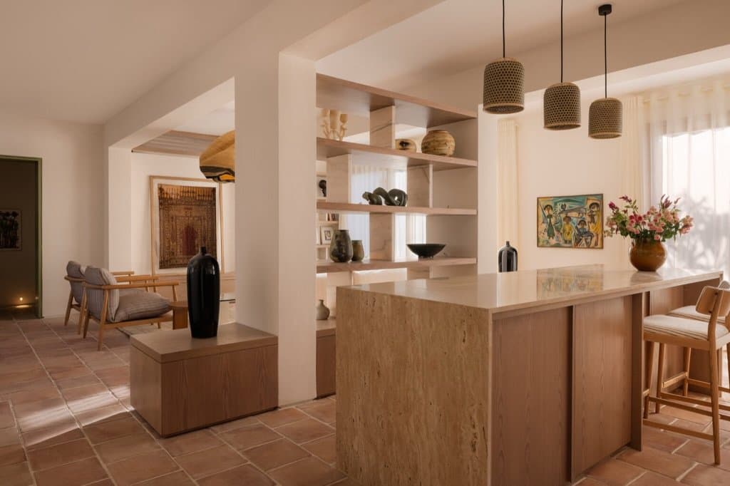 A travertine-clad island anchors the open-plan kitchen, its cane pendants and shelved ceramics quietly articulating the space.
