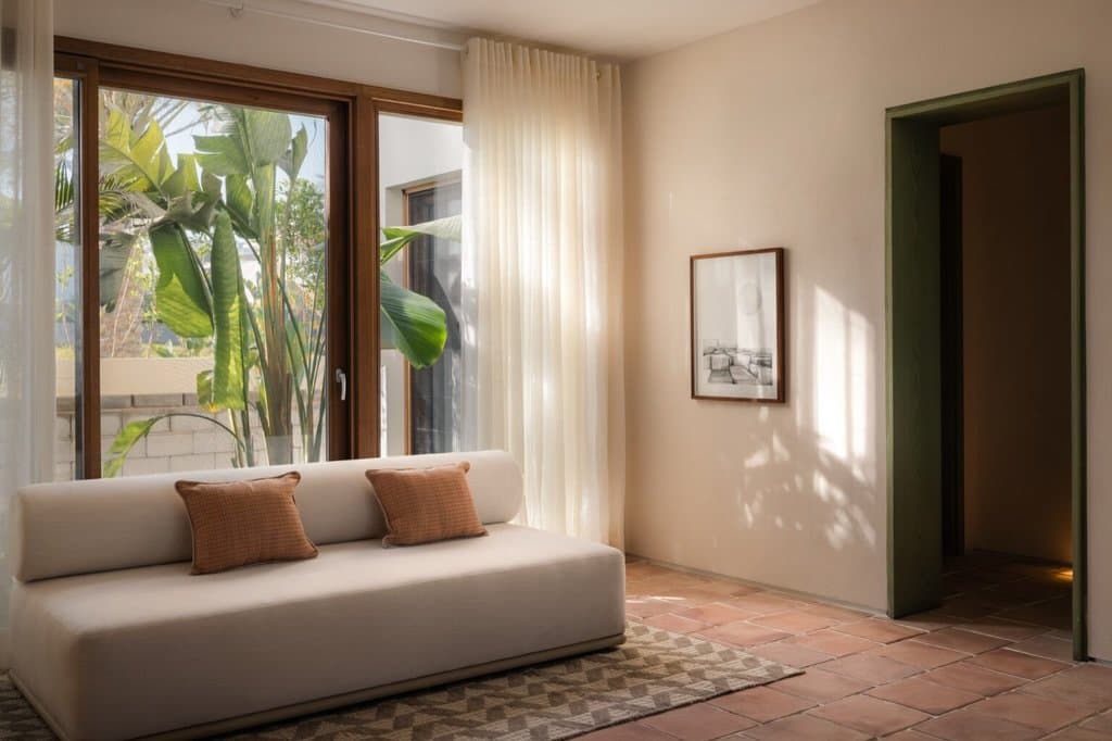 A daybed corner beside a timber-framed window, where sheer light and a green-painted doorway introduce a note of memory.