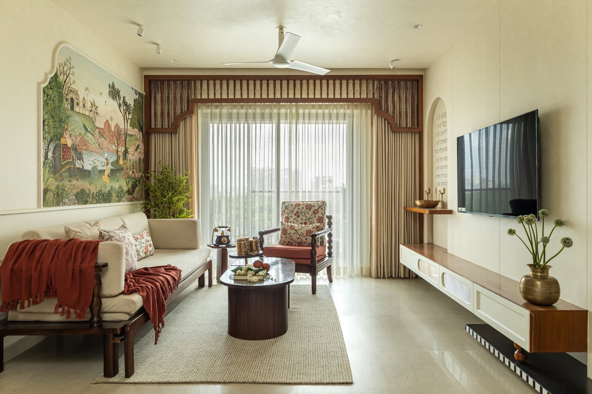 The living room, where a Mughal miniature-inspired mural anchors the seating composition and a carved jharokha-style valance frames the balcony